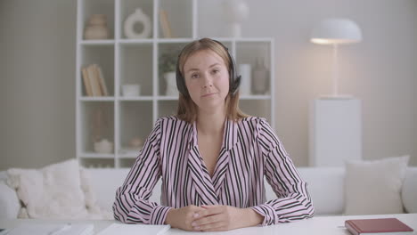young-friendly-woman-is-listening-interlocutor-by-video-chat-by-headphones-and-nodding-looking-at-camera